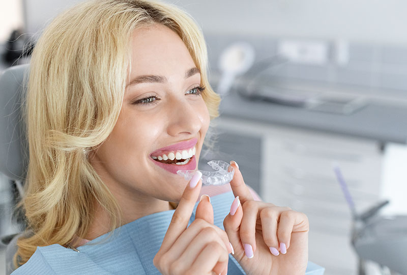 A smiling woman wearing dental braces, holding them up with both hands while seated at a dental chair.