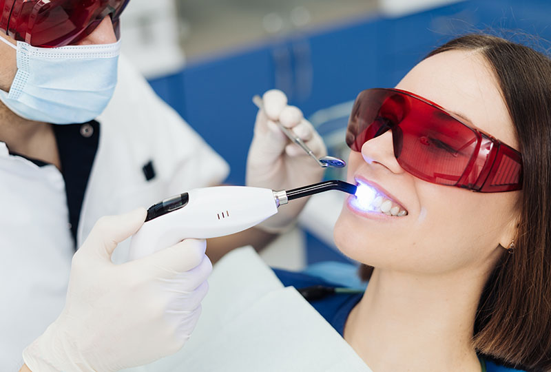 The image shows a dental professional performing a dental procedure on a patient who is wearing red protective eyewear.
